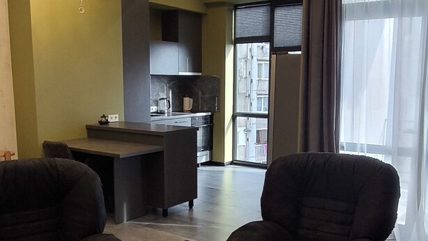 A modern living room with dark sofas, a wooden coffee table, and a kitchen area in the background.