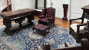 A room with a large wooden table, a red armchair, and a matching ottoman. There is a patterned rug on the floor, a tall mirror, and a wooden cabinet.