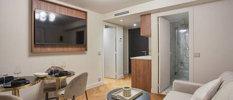 Una sala de estar moderna con un sofá, una mesa de comedor con sillas, un televisor montado en la pared y una cocina visible a través de una puerta abierta.