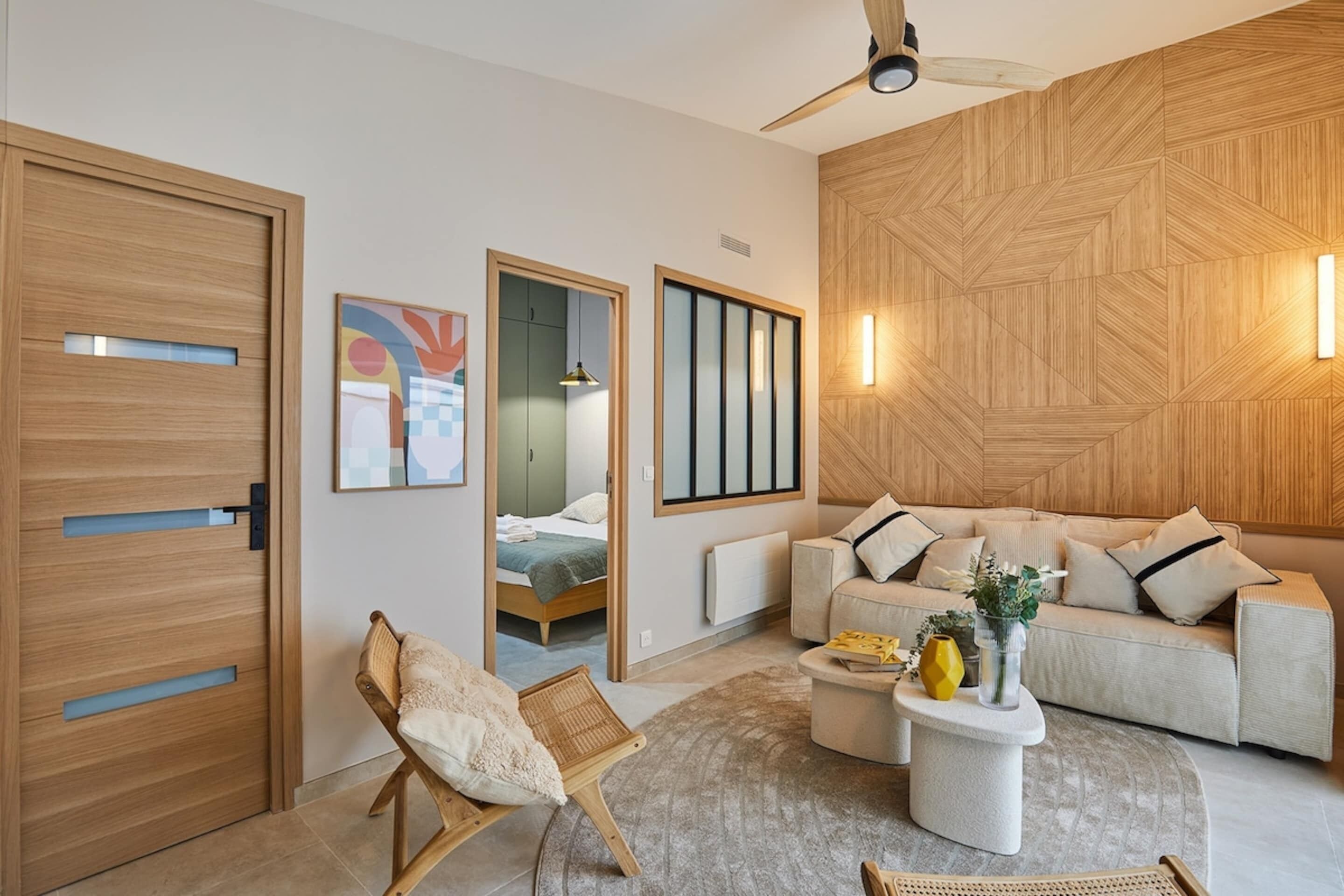 Un salon moderne avec un canapé, une table basse et un fauteuil. On y trouve une porte en bois et un mur orné d’un motif géométrique.