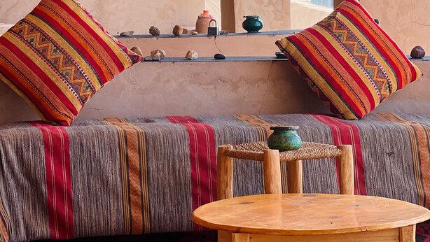 A traditional room with a wooden ceiling, a sofa with patterned cushions, a small table, and a view through a window. - Camp Mbark authentic (M'Hamid El Ghizlane)