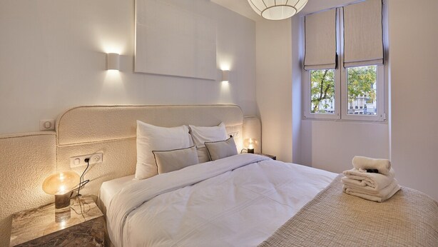 A modern bedroom with a large bed, two bedside lamps, a nightstand, and a window with curtains. - Superb Apartment in the Heart of Paris (Paris)