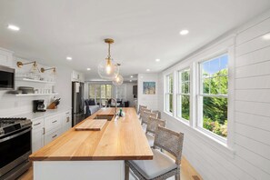 A modern kitchen with a long wooden island, stainless steel appliances, and large windows offering a view of greenery.