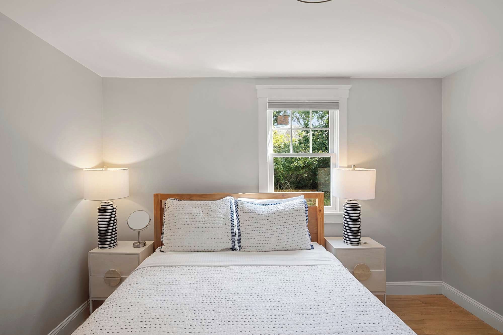 A bedroom with a large bed, two bedside tables, a window with a view of greenery, and a ceiling light.