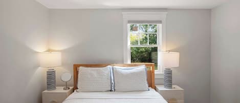 A bedroom with a large bed, two bedside tables, a window with a view of greenery, and a ceiling light.