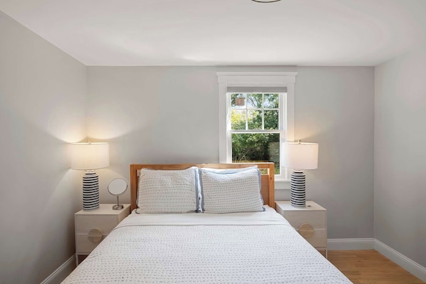 A bedroom with a large bed, two bedside tables, a window with a view of greenery, and a ceiling light.