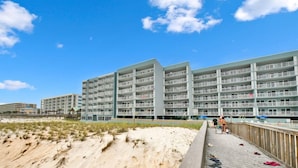 Um hotel na praia com vários andares, uma passarela de madeira e uma área de praia arenosa.