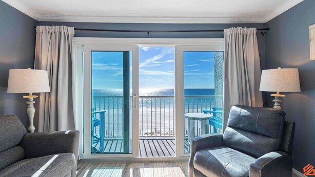 Quarto com vista para a praia através de grandes portas de vidro, uma pequena mesa redonda, duas cadeiras e duas luminárias.