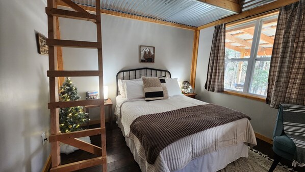 Un dormitorio con una escalera de madera, una cama con ropa de cama blanca, un pequeño árbol de Navidad y una ventana con cortinas.