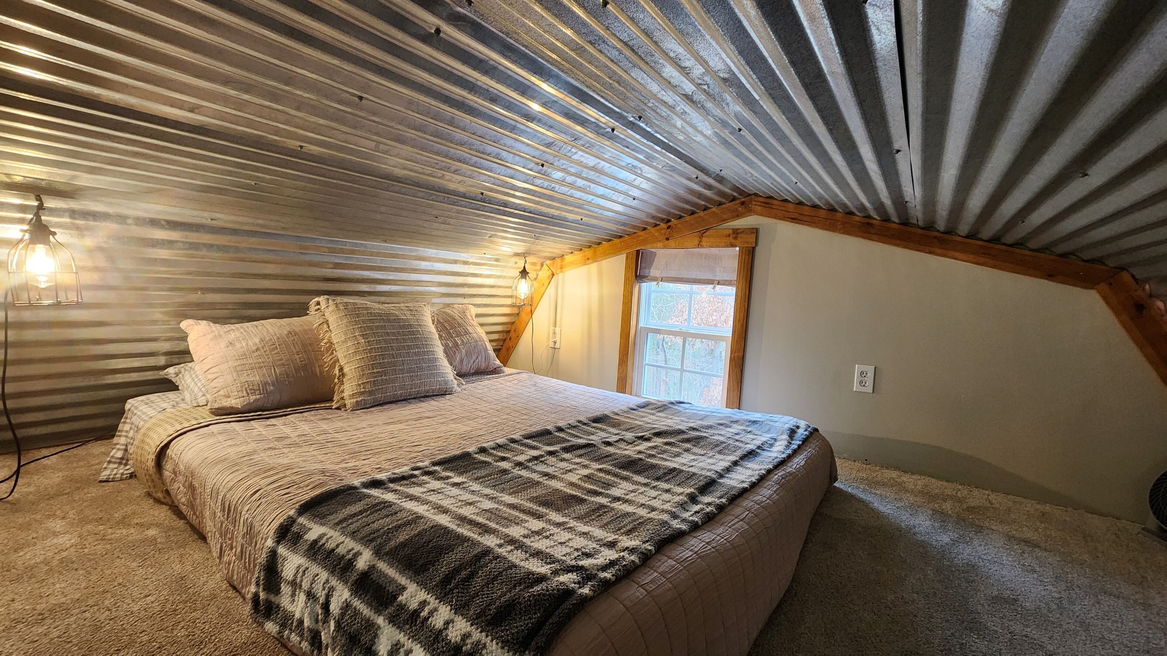 A bedroom with a sloped ceiling, a bed with a plaid blanket, and a window with a view of trees.