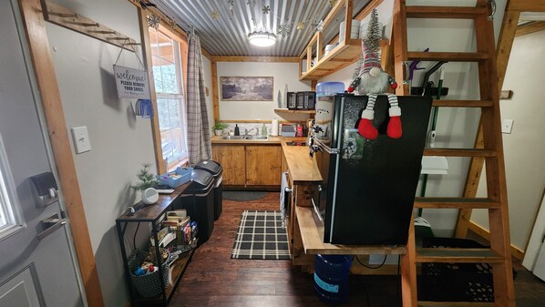 A compact kitchen with wooden cabinets, a black refrigerator, a microwave, and a sink. There is a small window with curtains, a welcome sign, and a ladder against the wall. - Cozy cabin located on mountainside hobby farm (Jasper)
