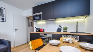 A modern kitchen with dark cabinets, a microwave, and a built-in oven. There is a dining table with white plates and wine glasses, and a chair with a yellow cushion. - Welcoming Apartment - 1br/4p - Arc de Triomphe (Paris)
