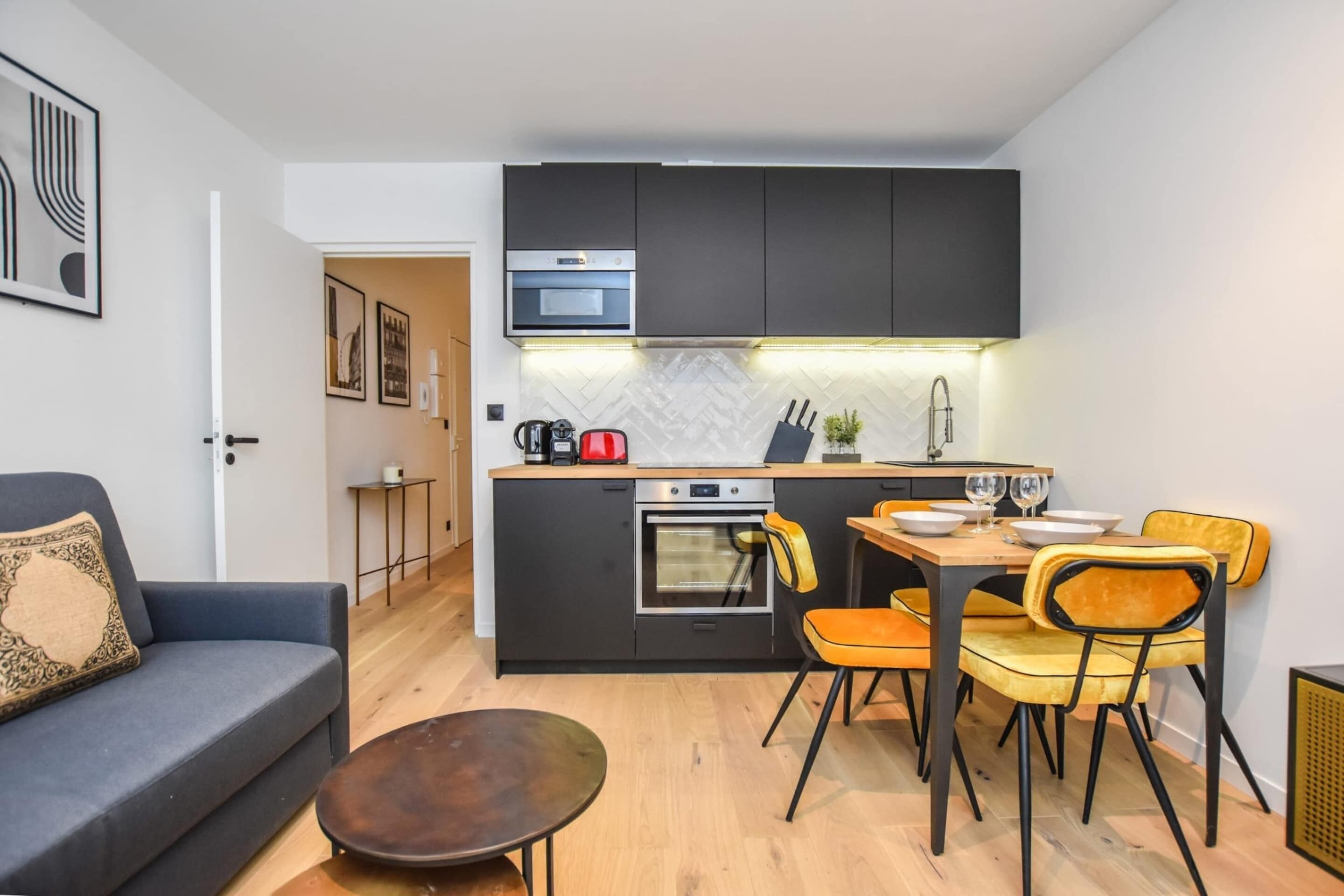 A compact kitchen with dark cabinets, a built-in oven, and a dining area with yellow chairs.