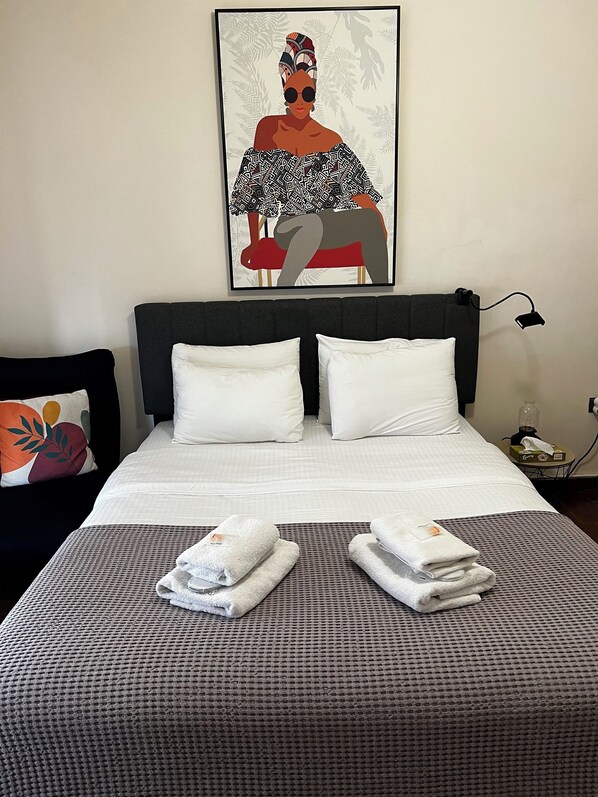 A neatly made bed with white linens, two pillows, and a patterned bedspread. There is a framed artwork above the bed, a bedside table with a lamp, and a wall-mounted phone. - Pnyka Apartment 3 (Athens)
