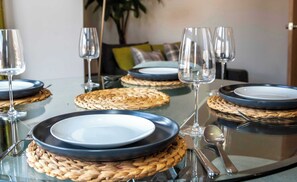 Mesa de jantar posta com pratos, copos e talheres, com jogos americanos trançados e tampo de vidro.