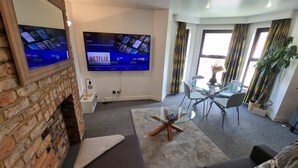 A living room with a brick wall, a flat-screen TV displaying Netflix, a glass coffee table, a sofa, and a dining area with a glass table and chairs.