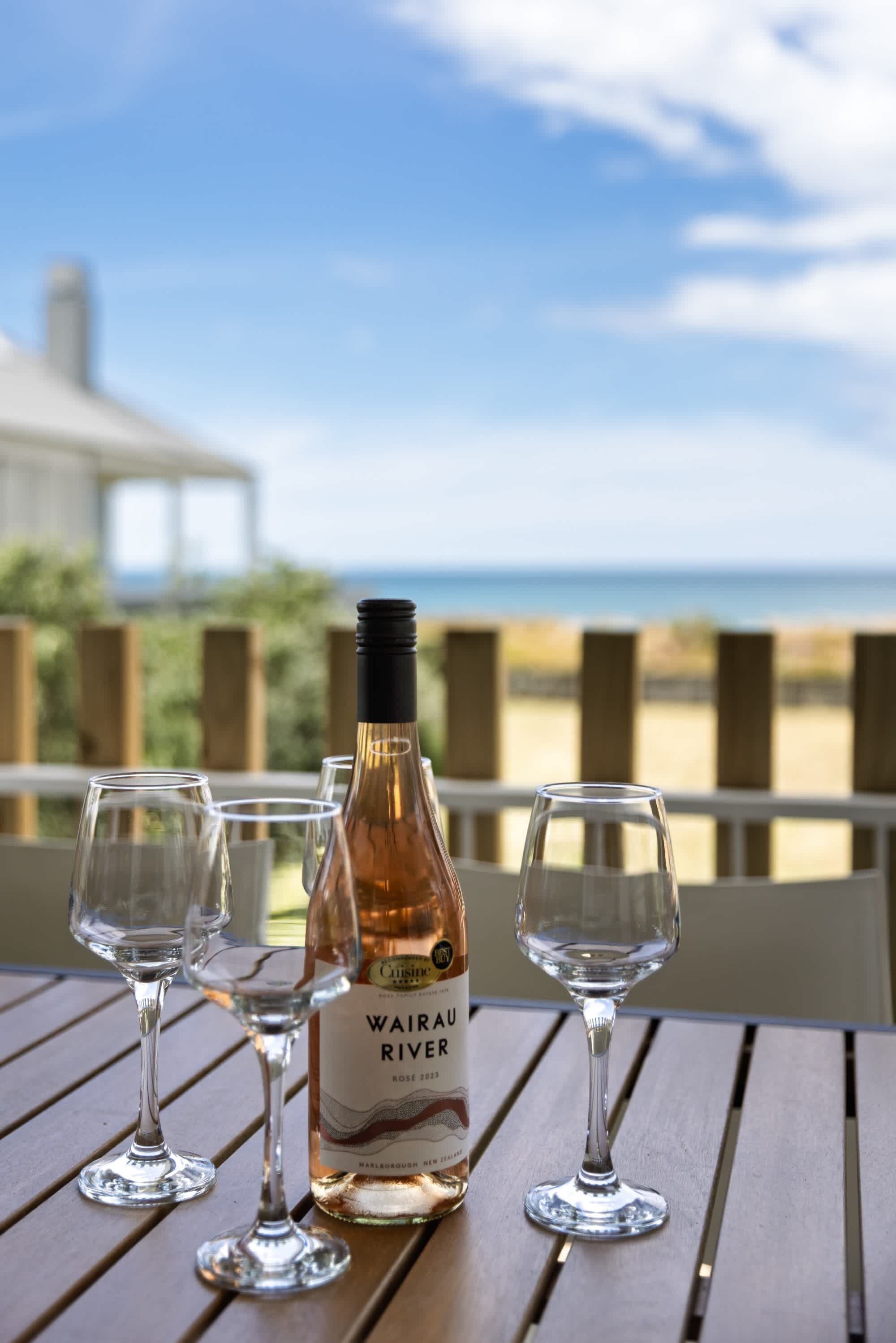 A bottle of Wairau River wine and three glasses on a wooden table with a view of the ocean and a wooden fence in the background.