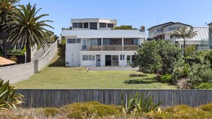 A two-story house with a flat roof, a balcony on the upper floor, and a fenced yard with grass and shrubs.