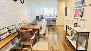 A spacious dining area with a long wooden table, wicker chairs, and a bench with striped cushions. A kitchen is visible in the background with a modern design. - Beautiful townhome with community pool, shuttle to Bethany, and more! (Millville)