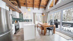 A modern kitchen with a large island, wooden ceiling beams, and large windows offering a view of a snowy landscape. - Spa, Steam room and Lake | Le Stuga (Lac-aux-Sables)