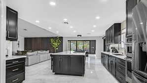 A modern kitchen with dark cabinetry, a central island, and a large refrigerator. - Luxury 6BR Vegas Villa w Pool &Spa, Near The Strip (Las Vegas)
