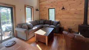 Uma sala de estar aconchegante com um sofá seccional, uma mesa de centro de madeira e uma lareira. Há duas janelas que oferecem luz natural e vista para o exterior.
