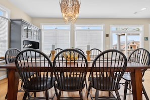 A dining area with a wooden table and chairs, a chandelier, and a view through large windows.