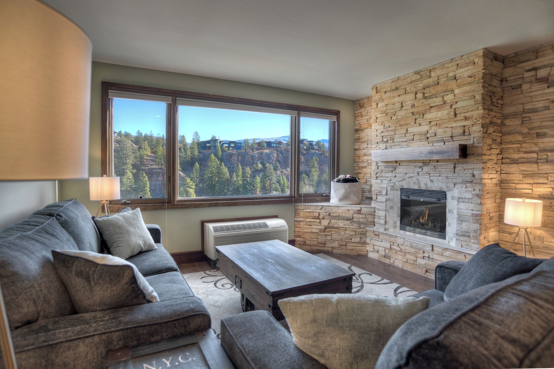 A living room with a stone fireplace, large windows, and a view of a forested area.