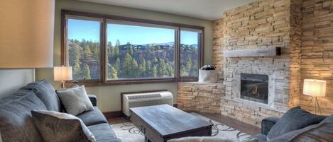 A living room with a stone fireplace, large windows, and a view of a forested area.