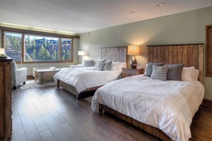 A bedroom with two beds, a nightstand, a lamp, and a view of the forest through large windows. - Down Home in Durango | AC, Pool, Hot Tub, Golf & Ski (Durango)