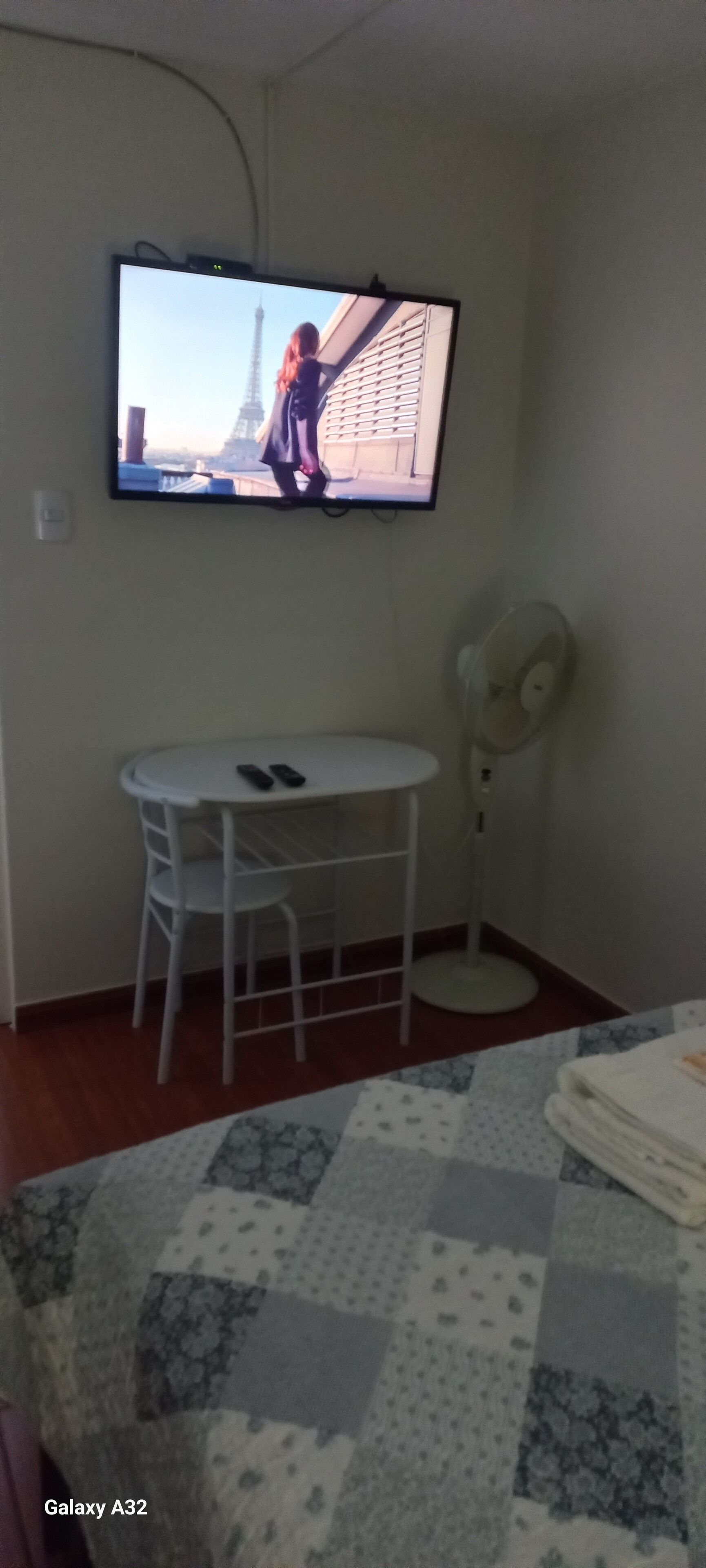Ein Hotelzimmer mit an der Wand montiertem Fernseher, einem kleinen runden Tisch mit Stühlen, einem Standventilator und einem Bett mit einer Steppdecke.