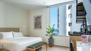 Habitación de hotel con cama, escritorio con computadora, planta en maceta y ventana con vista a un edificio.