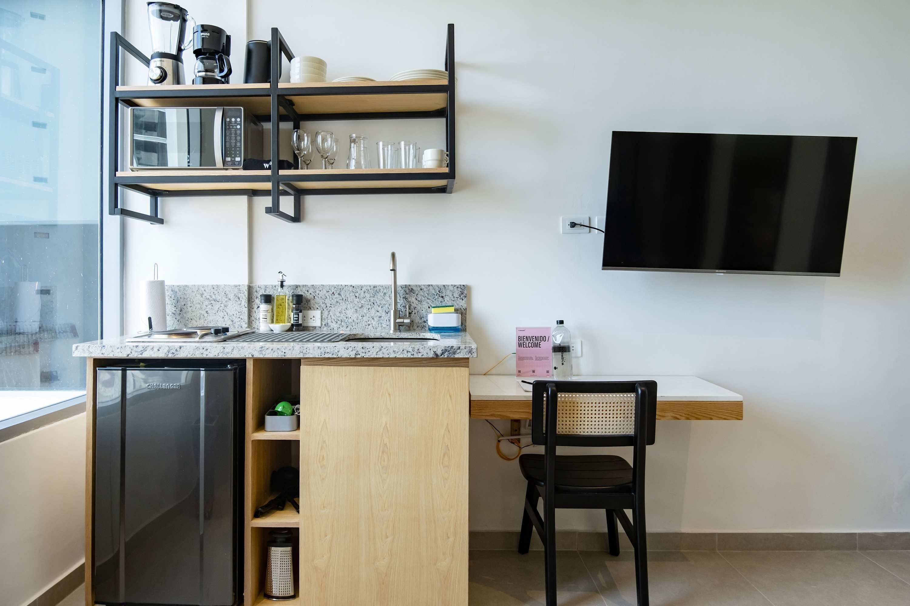 A compact kitchen with a built-in refrigerator, a microwave, and a coffee maker on a shelf. There's a wooden desk with a chair and a flat-screen TV mounted on the wall.
