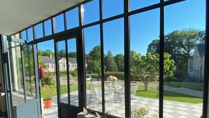Una habitación con ventanas amplias, suelo de baldosas y vistas a un jardín y edificios.