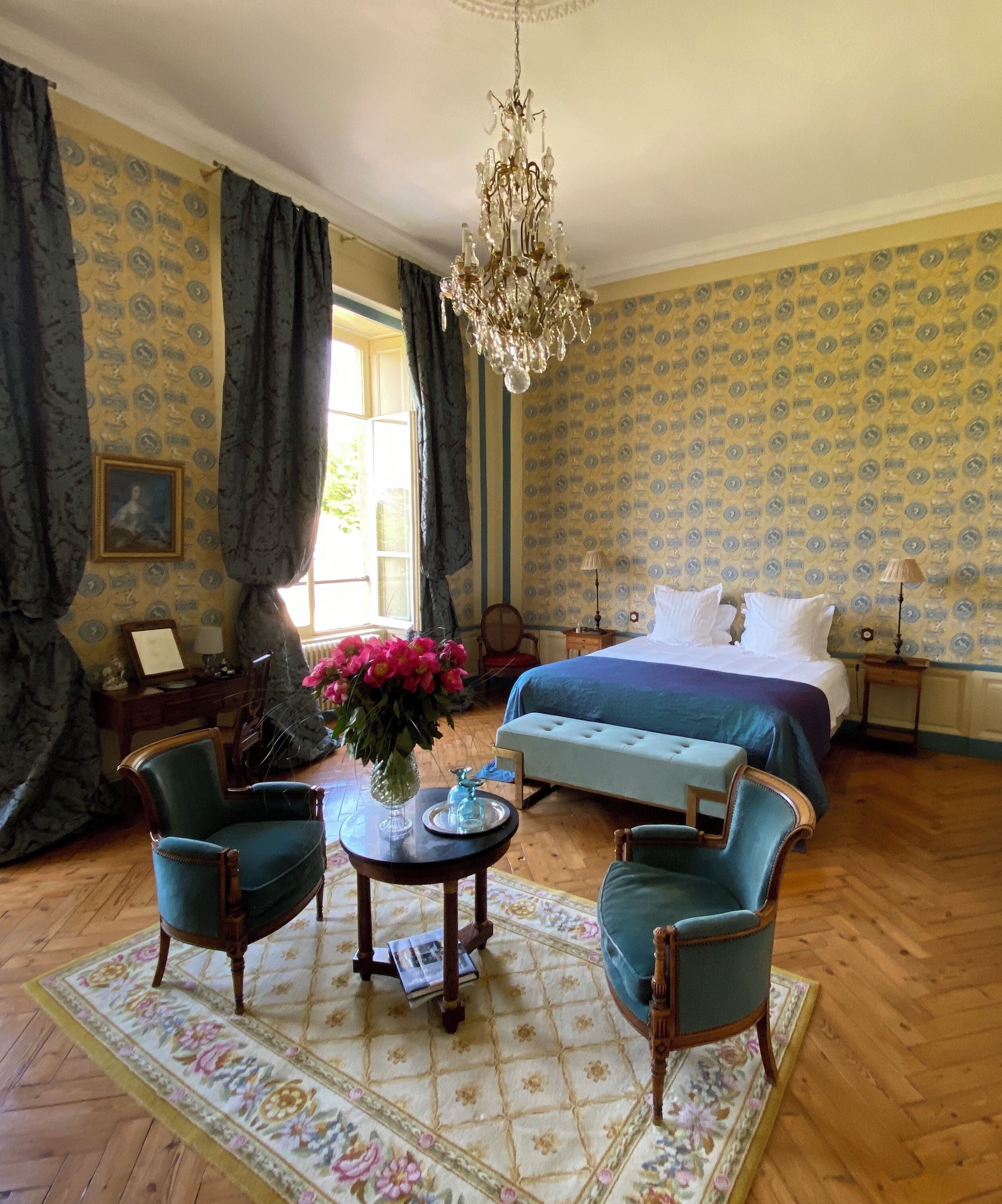 Slaapkamer met een groot bed, twee fauteuils, een klein tafeltje met een bloevas, een kroonluchter en behang met een patroon.