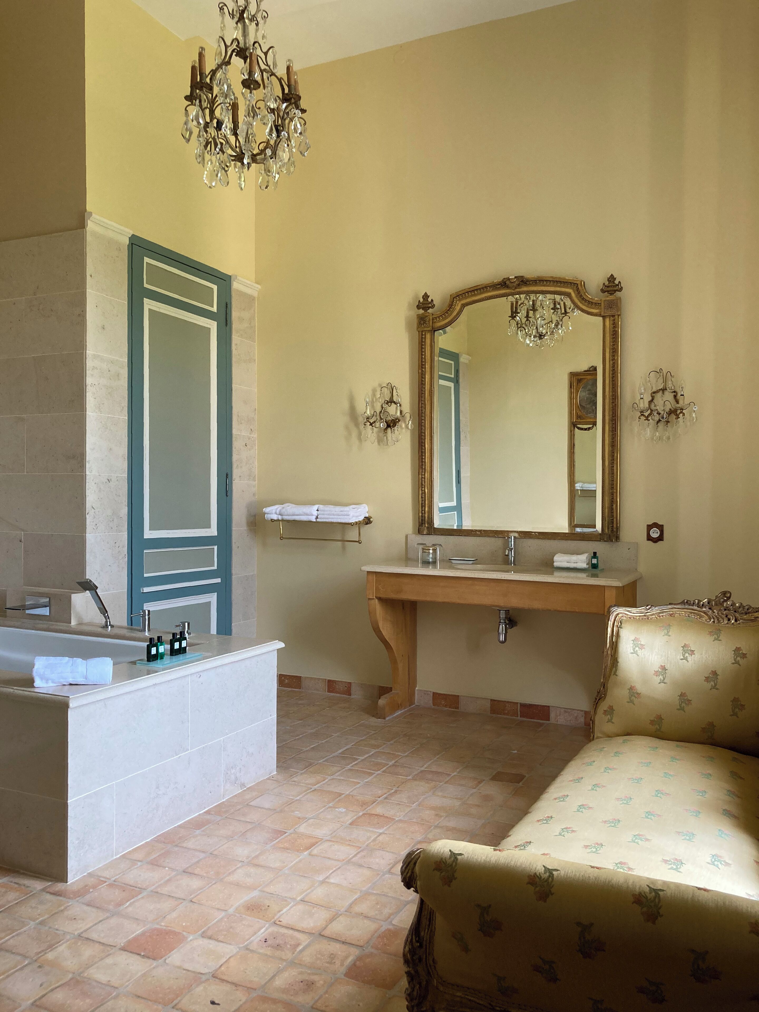 Un baño con bañera, tocador con espejo, una araña y una silla con estampado floral.