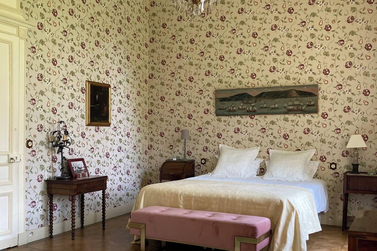 Un dormitorio con papel tapiz floral, una cama, un banco, una mesa y una araña.