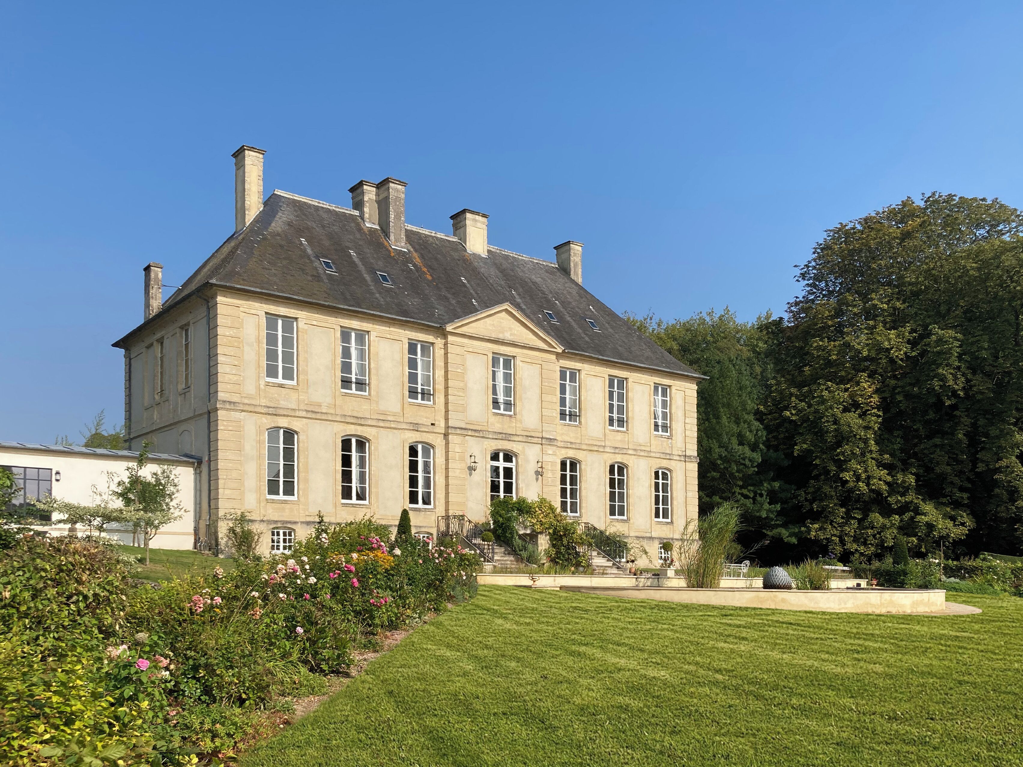 Ein großes, historisches Gebäude mit zahlreichen Fenstern und einer symmetrischen Fassade, umgeben von einem gepflegten Garten und blauem Himmel.