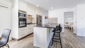 A modern kitchen with white cabinetry, a large island with bar stools, a microwave, oven, and refrigerator. - Bayview • Pet-Friendly Beach House for 6 • Fenced Yard • Walk to Beach (Agnes Water)