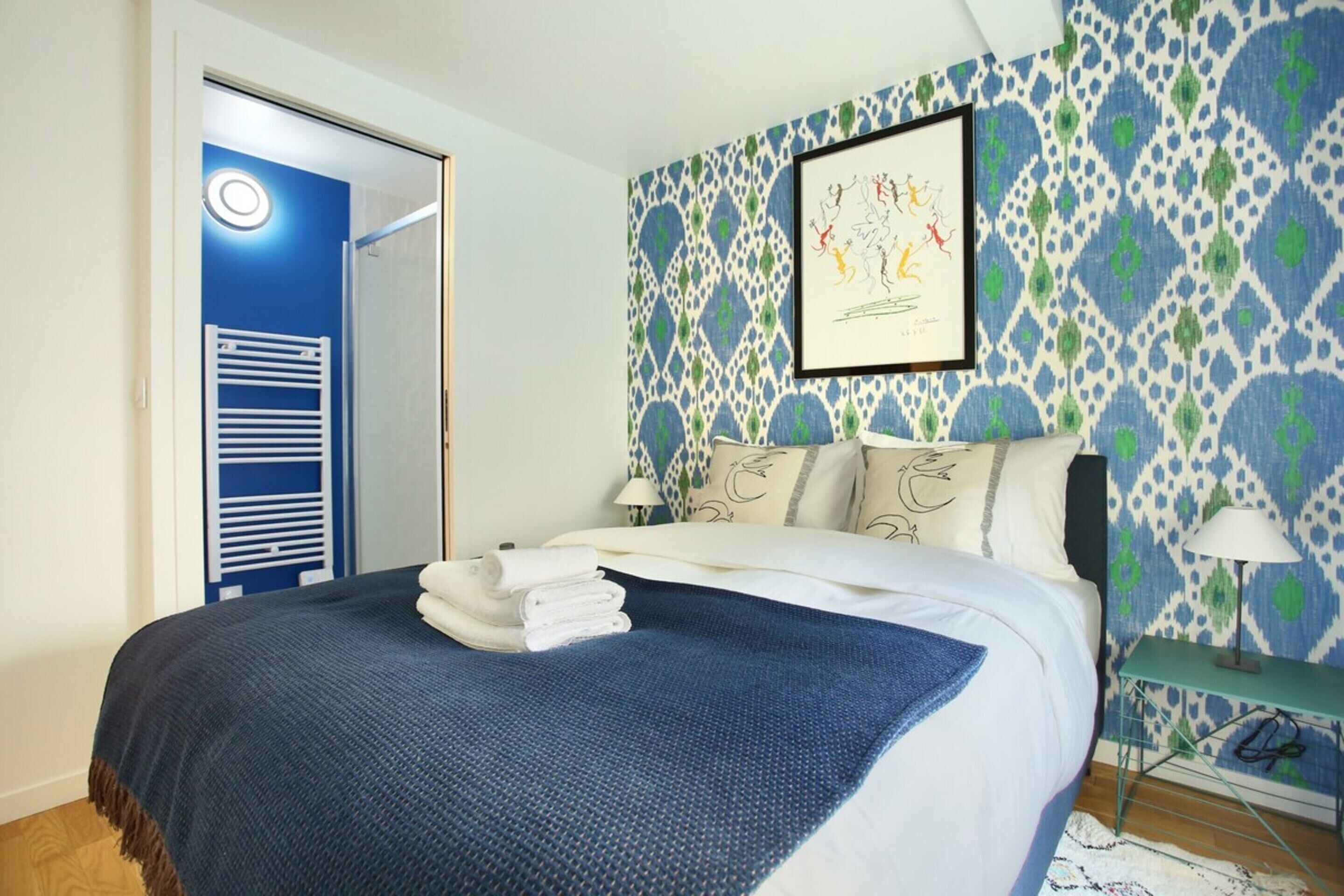 Un lit bien fait, avec une couverture bleue et des serviettes blanches, un mur décoré d’un motif, une œuvre d’art encadrée et une table de chevet avec une lampe.