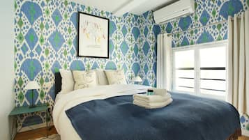 A bedroom with patterned wallpaper, a bed with a blue blanket, a framed artwork, and an air conditioning unit.
