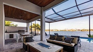 Cozinha externa moderna com bancada de pedra, banquetas e área de estar com sofá e almofadas.