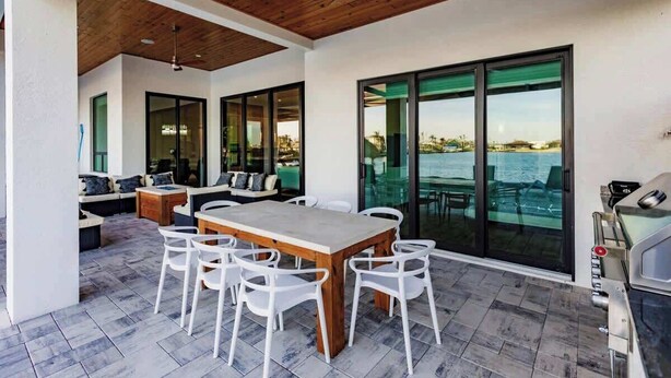 A modern outdoor patio with a wooden ceiling, large sliding glass doors, a dining table with chairs, and a barbecue grill. - Skyline Paradise Retreat (Cape Coral)
