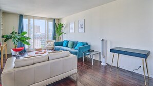Ein modernes Wohnzimmer mit einem weißen Sofa, einem blauen Sessel und einem blauen Sofa mit grünen Kissen. Zwei Blumentöpfe, ein kleiner Tisch mit einem Buch und eine Stehlampe sind ebenfalls zu sehen.