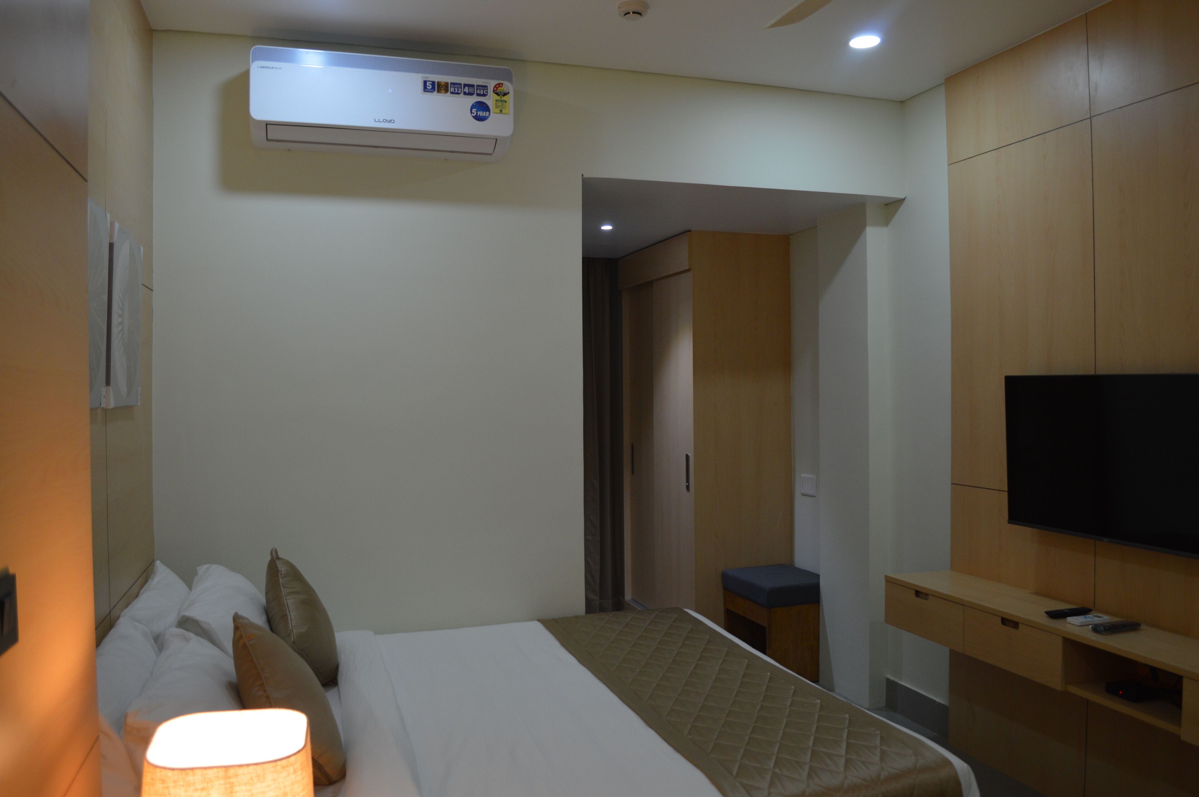 A hotel room with a bed, bedside lamp, TV, and an air conditioning unit.