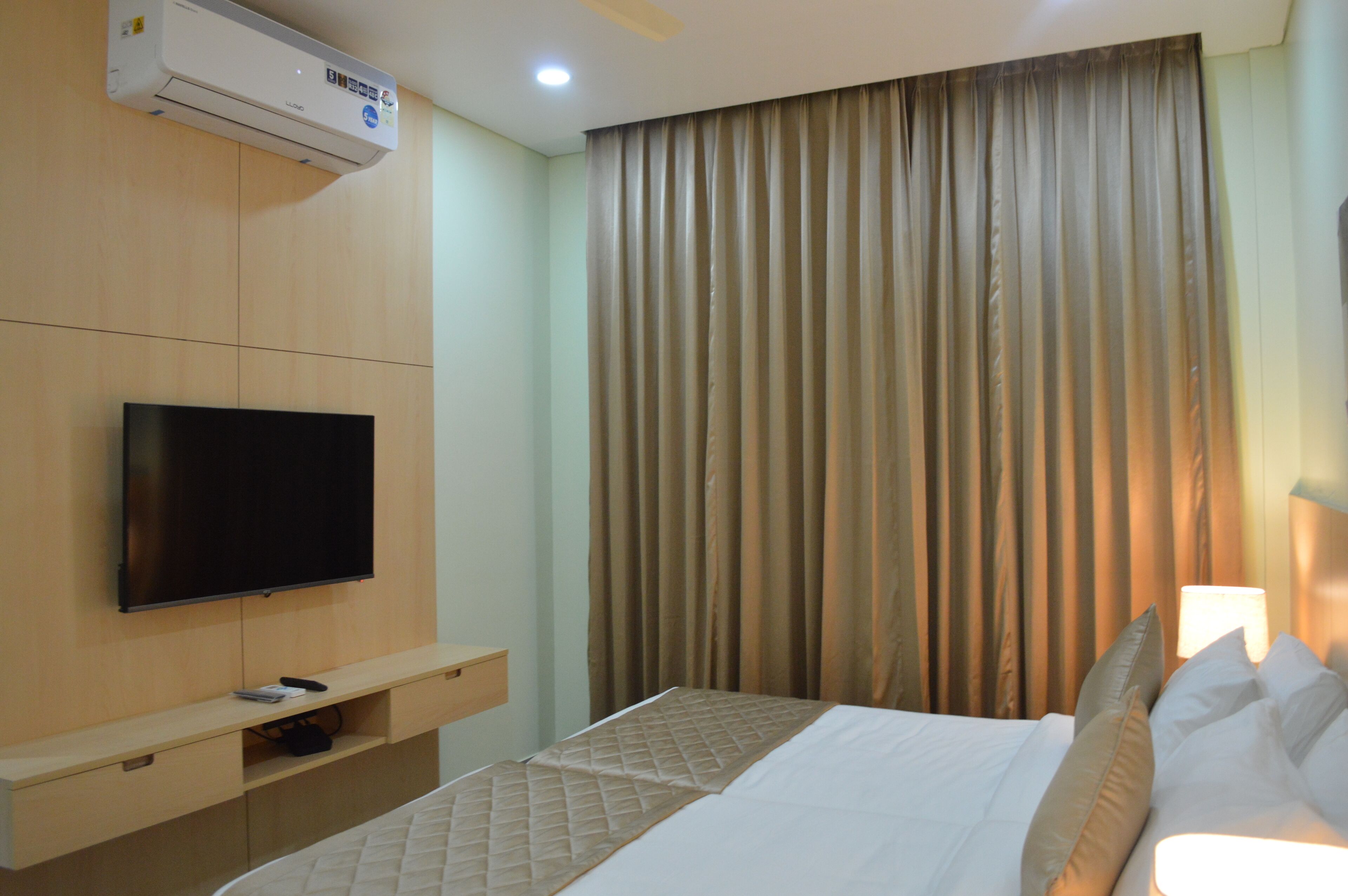 A hotel room with a flat-screen TV mounted on the wall, a bed with white bedding, a bedside table with a lamp, and a large window with curtains.