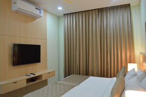 A hotel room with a flat-screen TV mounted on the wall, a bed with white bedding, a bedside table with a lamp, and a large window with curtains.