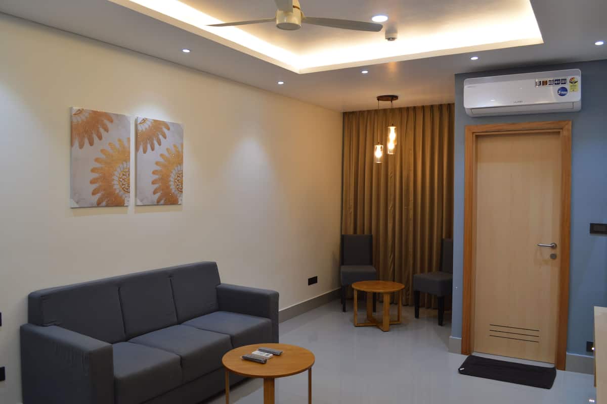 Moderni hotellihuone, jossa on harmaa sohva, kaksi pientä pöytää, kattotuuletin ja ovi, jonka yhteydessä on ilmastointilaite.