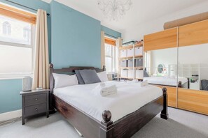 Un lit bien fait, avec du linge de lit blanc et une serviette pliée, une table de chevet avec une lampe, une armoire avec un miroir et une fenêtre avec des rideaux.
