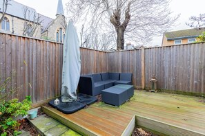 Une terrasse en bois dotée d’un espace salon intégré, d’un espace extérieur couvert et offrant une vue sur le clocher d’une église en arrière-plan.